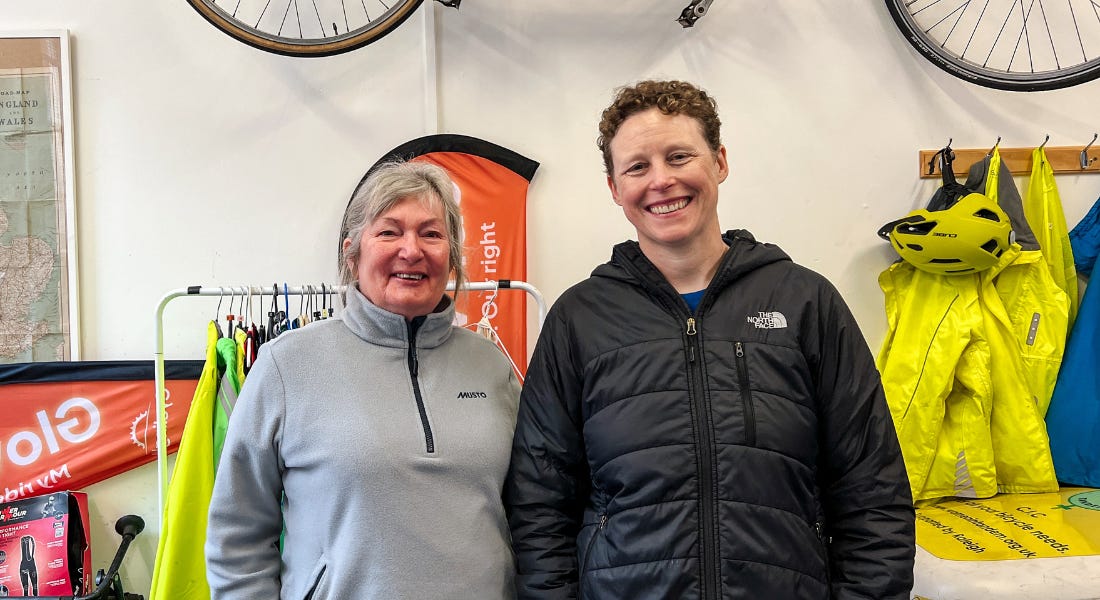 Two women wearing jackets stood in front of a jacket rail with a bike above them Two women wearing jackets stood in front of a jacket rail with a bike above them