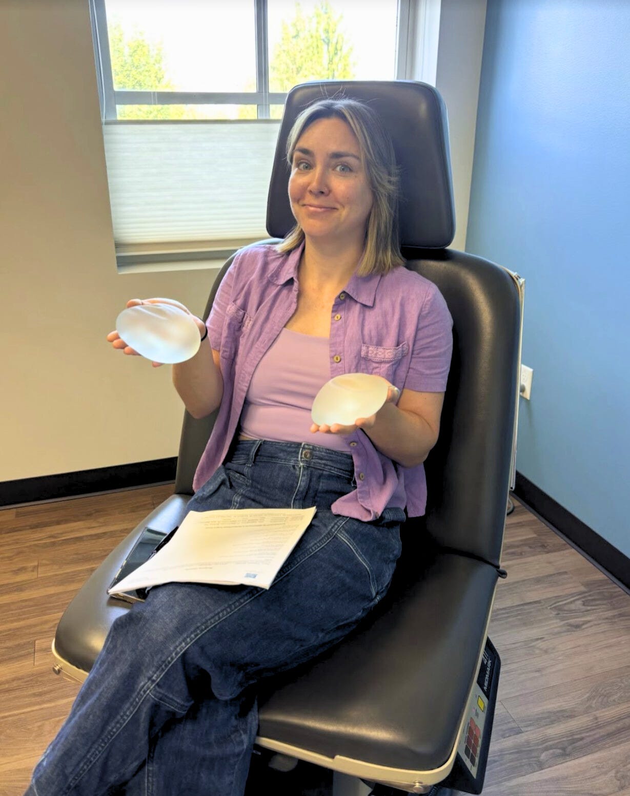 Cassidy sitting in a doctor's chair in the plastic surgeon's office, holding two breast implant options