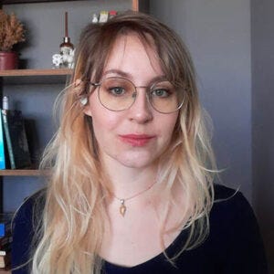 Bruna Orsi - Mulher de cabelos loiros compridos, olhos azuis, usando óculos de aros redondos, blusa preta e colar com pingente no pescoço, tendo ao fundo uma estante com livros e objetos decorativos