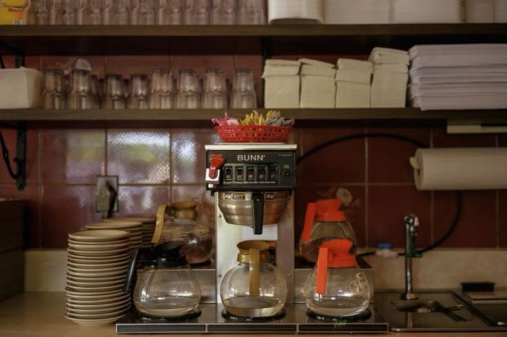 This may contain: a coffee maker sitting on top of a counter next to stacks of plates and cups