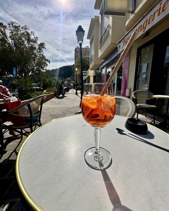 An Aperol spritz with a paper straw on a round café table on a sidewalk in Villefranche-sur-Mer, a boulangerie sign and a woman at the next table behind. An Aperol spritz with a paper straw on a round café table on a sidewalk in Villefranche-sur-Mer, a boulangerie sign and a woman at the next table behind.