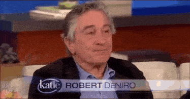 a man named robert deniro is sitting on a couch in front of a sign that says katie .