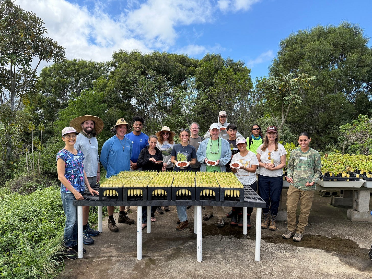 Group photo of fifteen people standing behind a table full of flats with seedlings. All different ages, from teens on up to seniors, and all shades of skin. A big wide smile on every face.