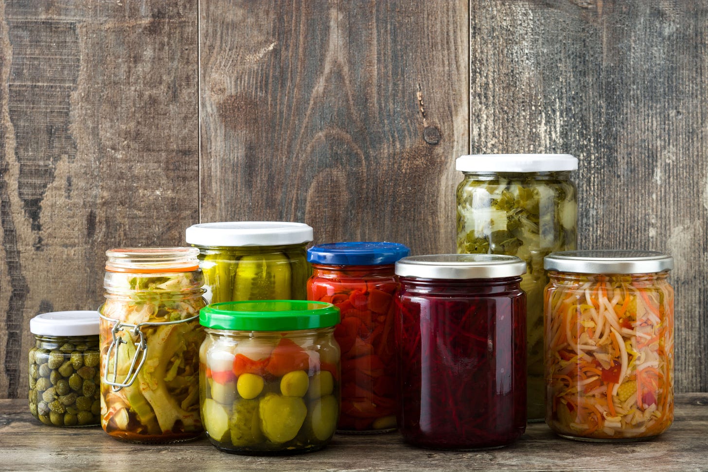 Several jars filled with preserved vegetables — cucumbers, olives, bell peppers, beets, and mixed vegetables — stand on a wooden table in front of a rustic wood wall.