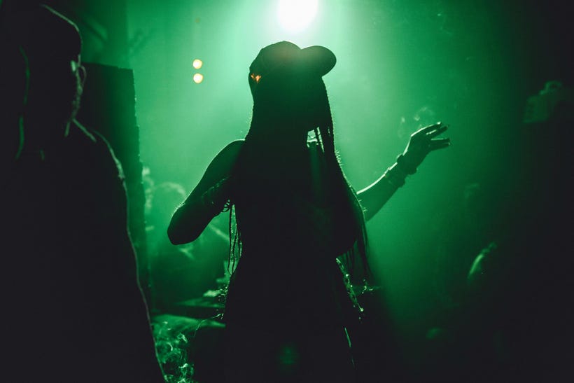 A woman with long thin braids and wearing a cap stands in silhouette under green lights in what looks like a nightclub. A woman with long thin braids and wearing a cap stands in silhouette under green lights in what looks like a nightclub.