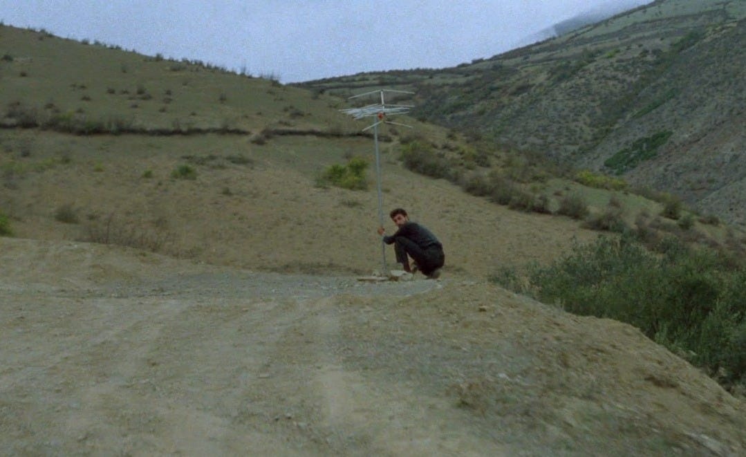 captura de tela do filme "e a vida continua", de abbas kiarostami. filmado da estrada, um moço olha em direção à câmera enquanto tenta ajustar a posição de uma antena na encosta do morro. ele está todo de preto