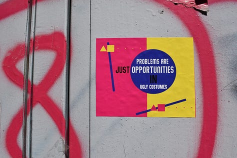 Gallery of 3 photos. #1- Side of gray shipping container with the number 8 with a large circle spray painted red. A sign in the circle reads: Problems are just opportunities in ugly costumes.” #2 A young woman helping an older woman walk on a busy city street. A large pink umbrella covers the women’s faces and most of their bodies as they walk along.  #3 Sharp corner of a grey stone building. A black sign with white letters curves around the corner of the building says: A change may be just around the corner.”