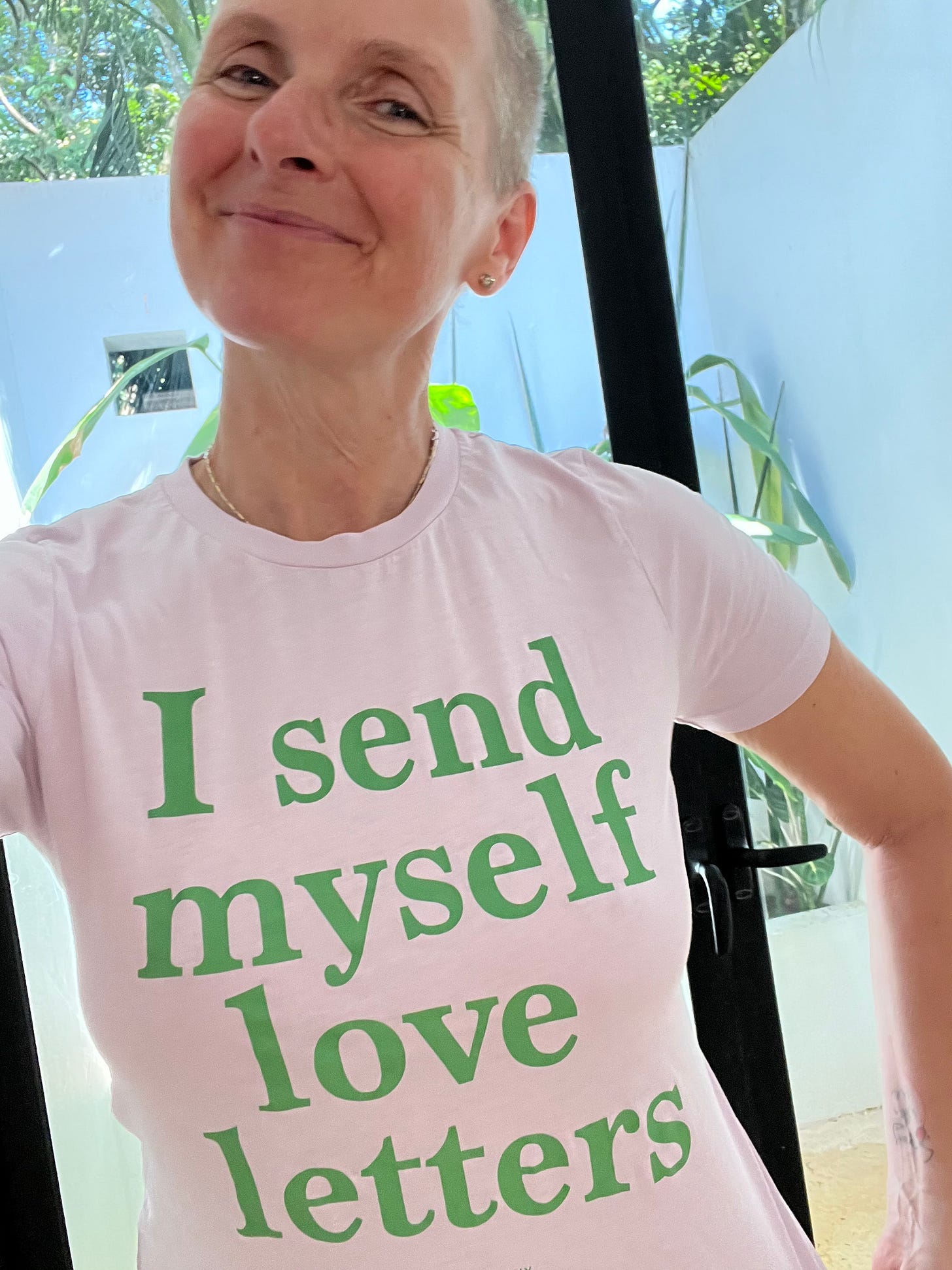 Photo of Elizabeth Gilbert. Gilbert is a white woman with buzzed gray hair. She is smiling and wearing a white t-shirt with green text on it. The green text reads, "I send myself love letters." Photo of Elizabeth Gilbert. Gilbert is a white woman with buzzed gray hair. She is smiling and wearing a white t-shirt with green text on it. The green text reads, "I send myself love letters."