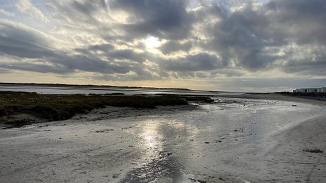 Six different shots of the beach with sand, rocks and shimmering water, several with blue skies, one with moody clouds and the sun peeking through.