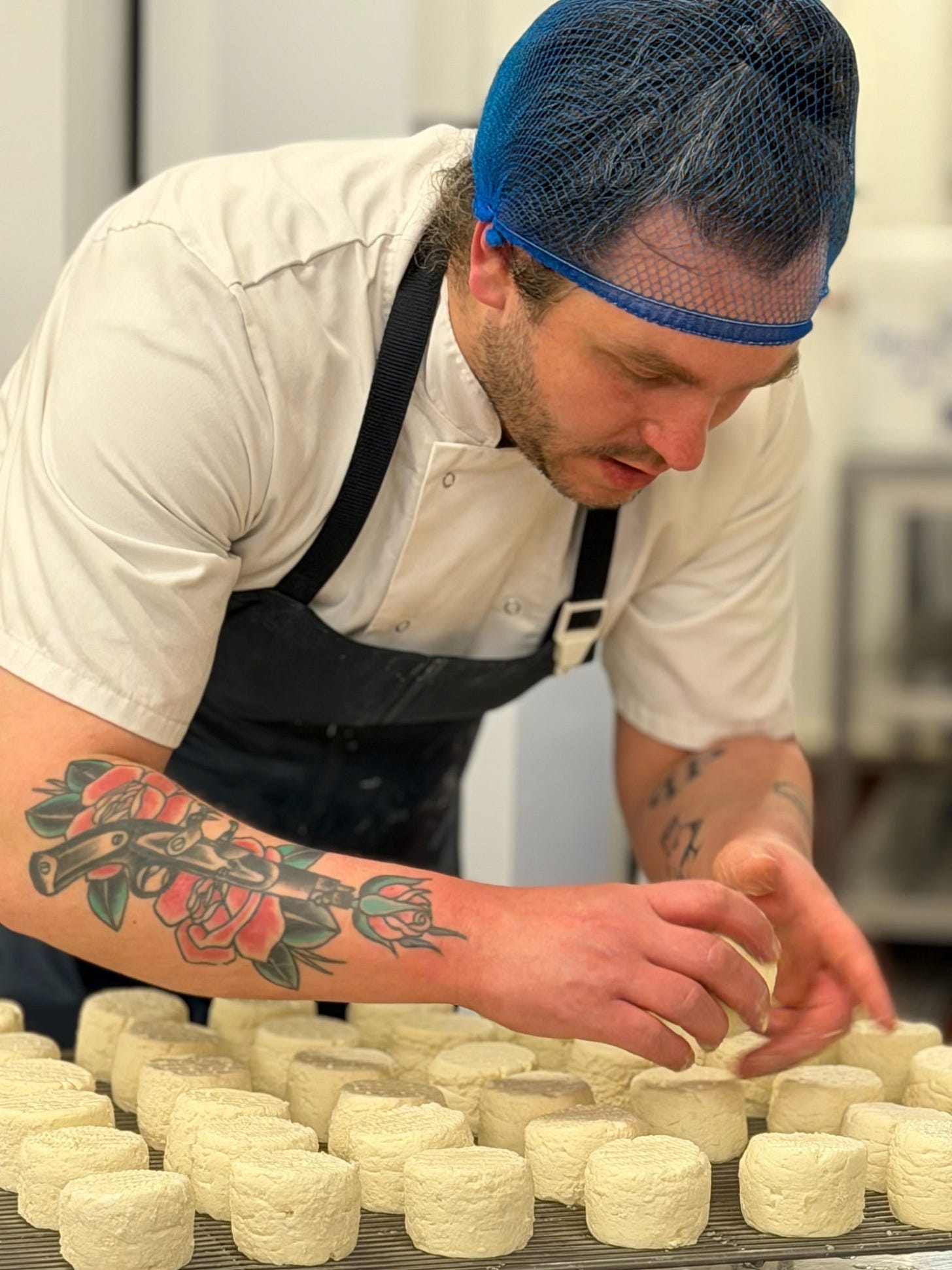 A man - Jake Goldstein - wearing an apron, chef's whites, and hair net leaning over a tray of cheese A man - Jake Goldstein - wearing an apron, chef's whites, and hair net leaning over a tray of cheese