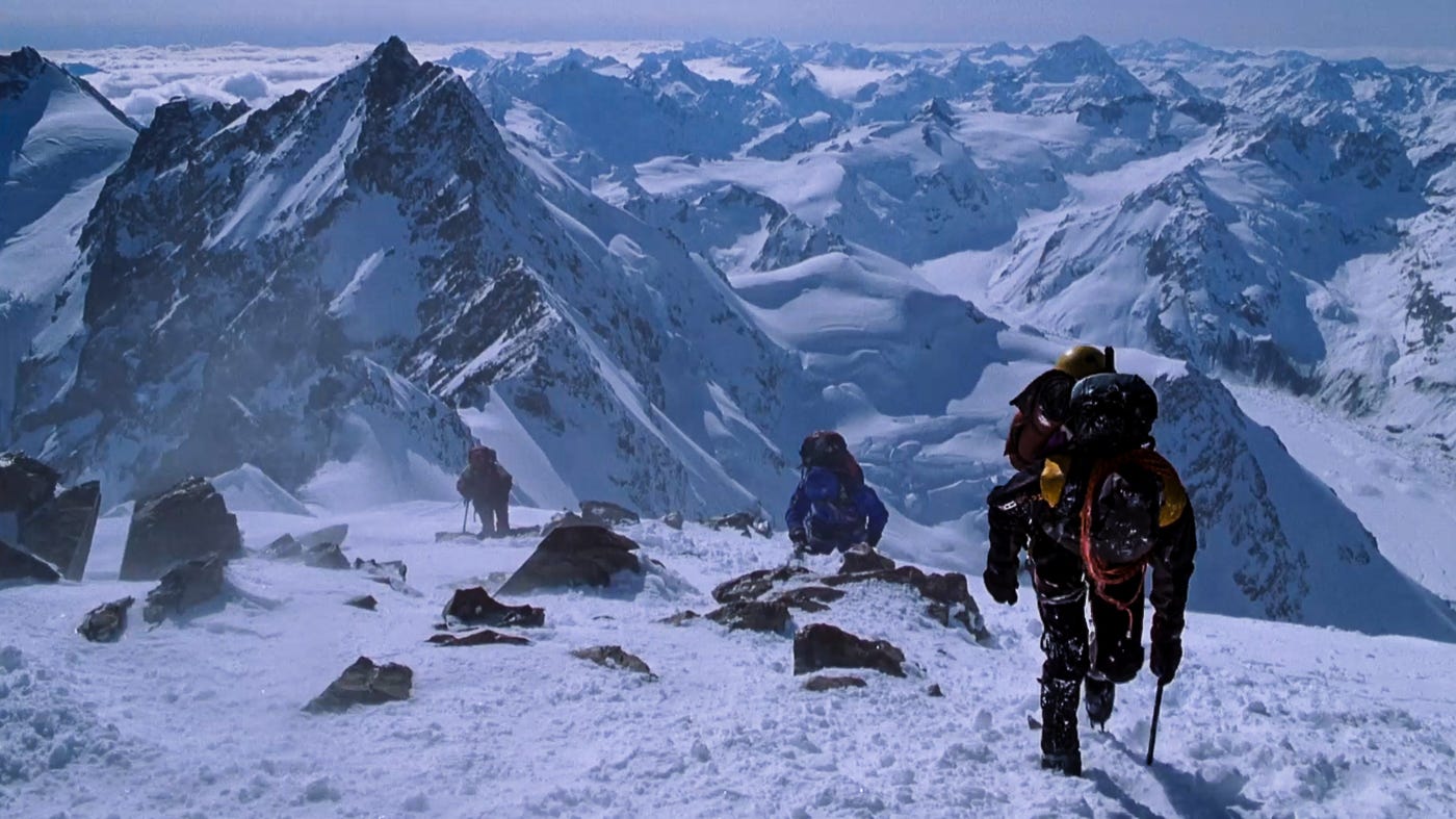 A team of climbers makes their final ascent toward the summit before an oncoming storm, surrounded by vast snow-covered peaks.