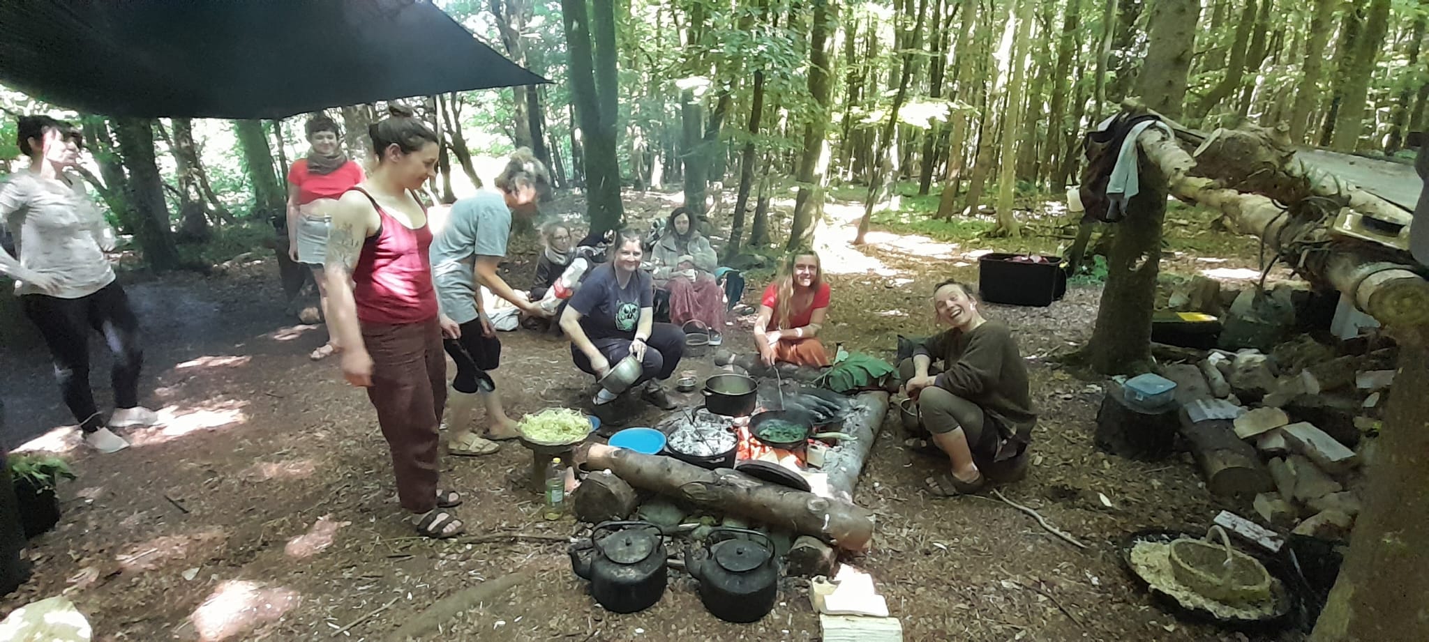 Communal gathering space around the forest hearth where 8 people are preparing food for our final feast, green woodland in view beyond them, Lucy O'Hagan is seated on the right of the picture, she is blonde and wearing earthy tones, behind here are some baskets and the wood pile.
