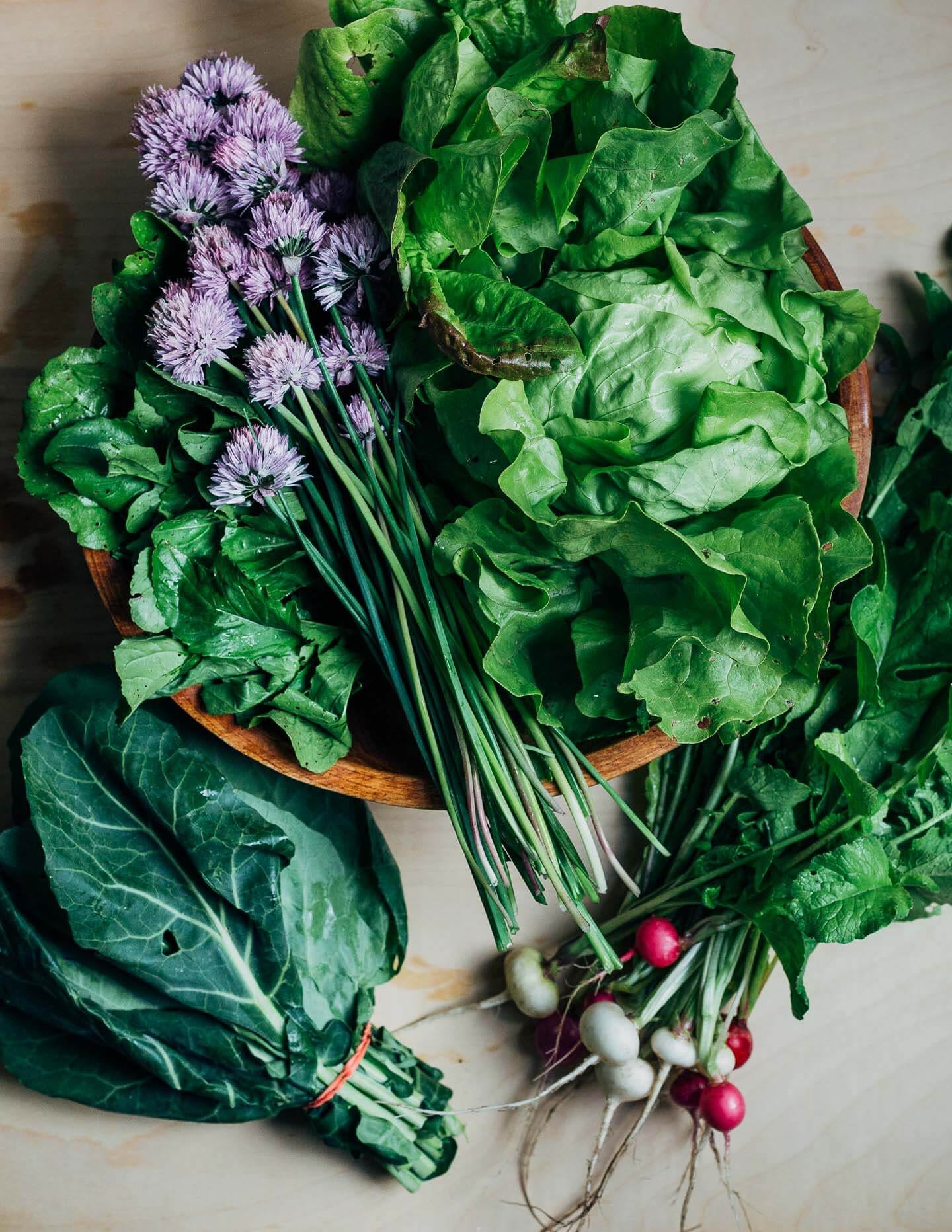 Lettuces, radishes, collards, and purple blooming chives arranged on a table. 