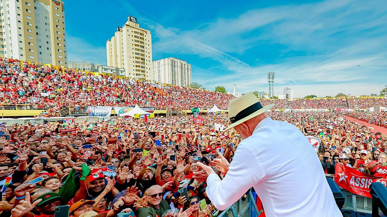 Lula, wearing a light-colored shirt and a straw hat, addresses a large crowd holding red flags at a campaign rally.