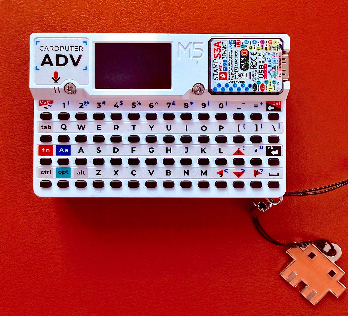 White M5Stack Cardputer ADV with a miniature keyboard and screen displayed on an orange table at Code with Claude 2026. White M5Stack Cardputer ADV with a miniature keyboard and screen displayed on an orange table at Code with Claude 2026.