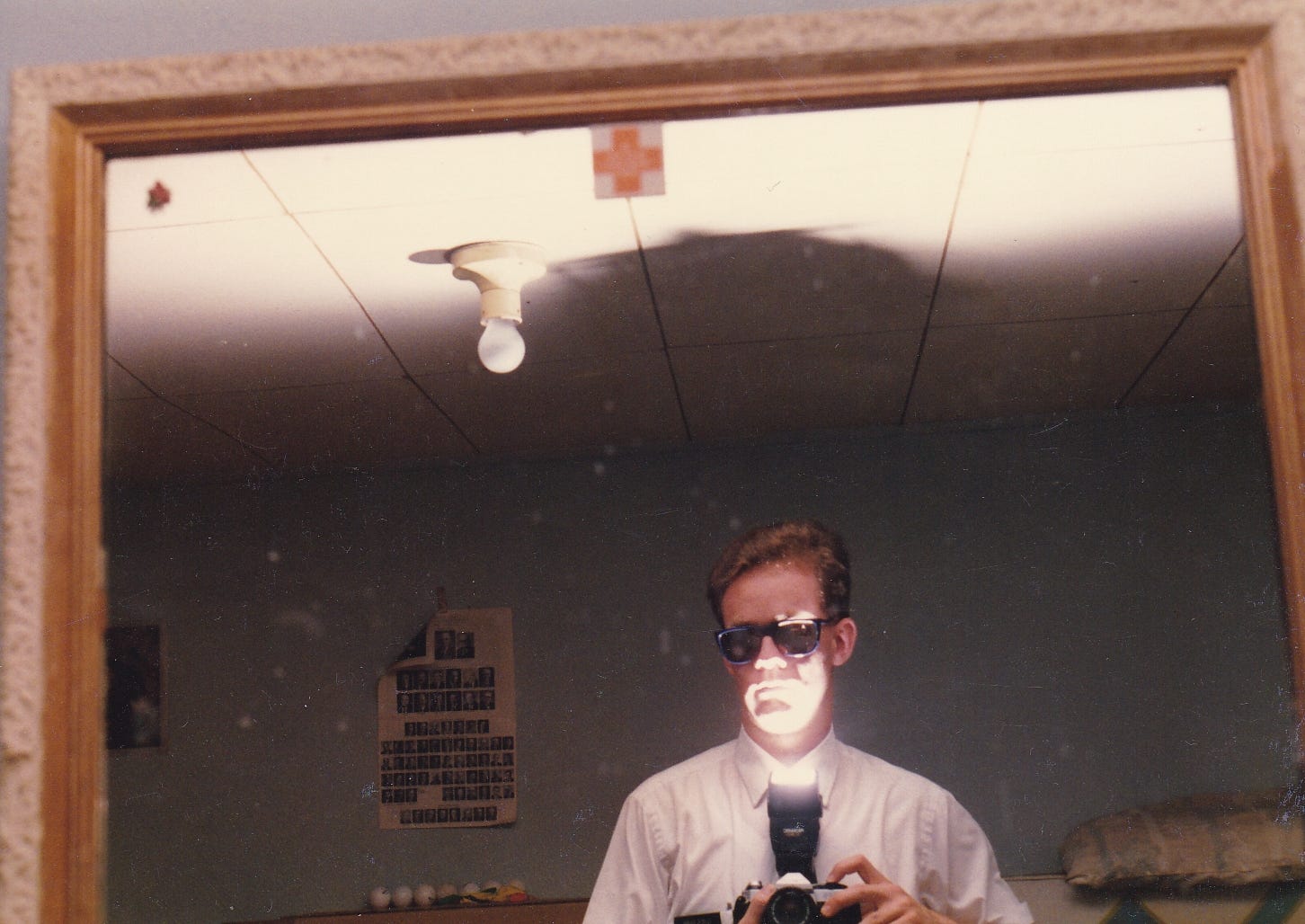 A young Mormon missionary wearing dark sunglasses looks in a mirror holding a camera in a dim room and takes a picture from chest level with the flash attachment pointed up at his face from below.