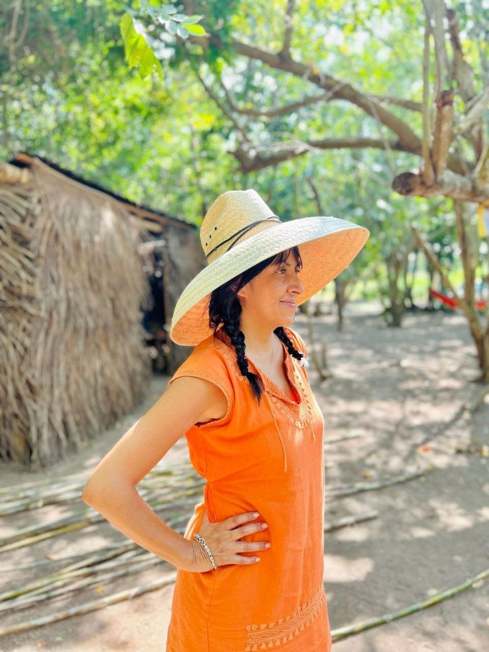 A woman in an orange embroidered dress and wide woven palm hat stands beside a palm-thatched hut