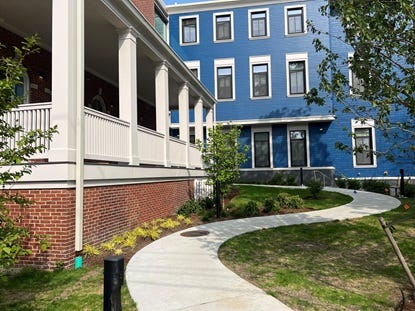 Sidewalk leading to a brick building with a covered porch (on left) and a three-story building painted blue. Sidewalk leading to a brick building with a covered porch (on left) and a three-story building painted blue.