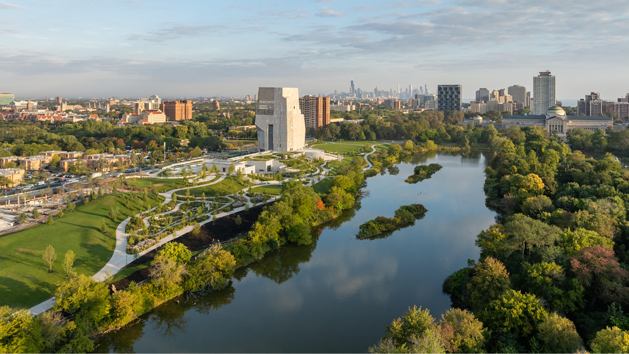 Aerial view of the Obama Presidential Center, a tall light stone museum tower rising above the landscaped grounds of Jackson Park in Chicago, with a lagoon and wooded islands in the foreground and the downtown skyline on the horizon. Aerial view of the Obama Presidential Center, a tall light stone museum tower rising above the landscaped grounds of Jackson Park in Chicago, with a lagoon and wooded islands in the foreground and the downtown skyline on the horizon.