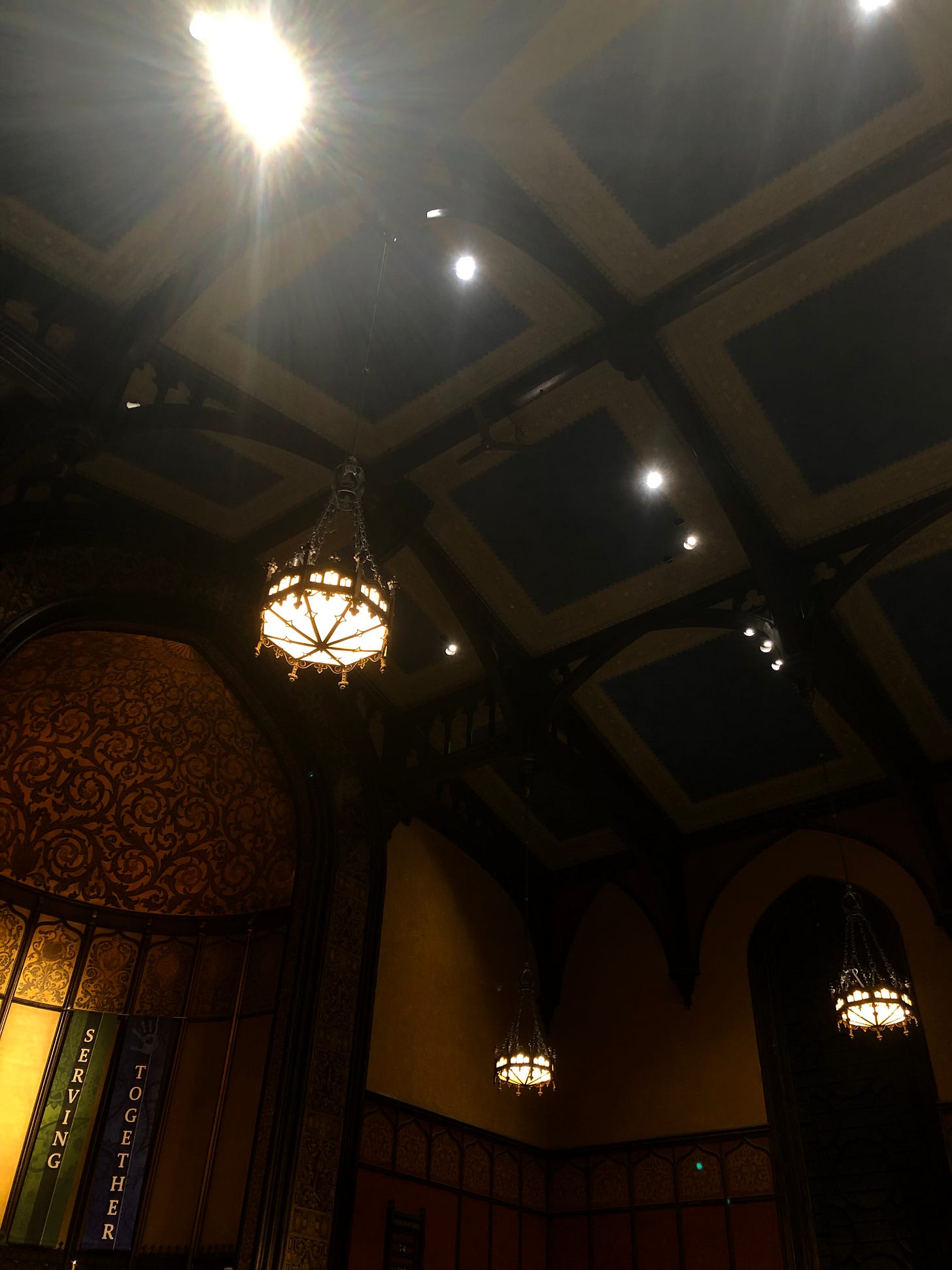 A dim, off-kilter photo of the ceiling of a church; two banners that read "serving" and "together" are visible in the bottom left corner