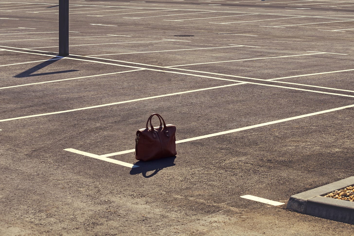 A brown leather briefcase sits upright in the middle of an empty asphalt parking lot, surrounded by painted white lines and long afternoon shadows. A brown leather briefcase sits upright in the middle of an empty asphalt parking lot, surrounded by painted white lines and long afternoon shadows.