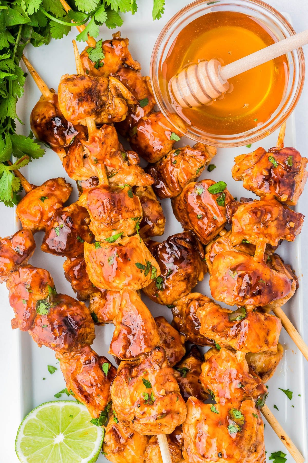 Grilled chicken skewers glazed with sauce, served with a bowl of honey and a honey dipper, garnished with fresh cilantro and a slice of lime.