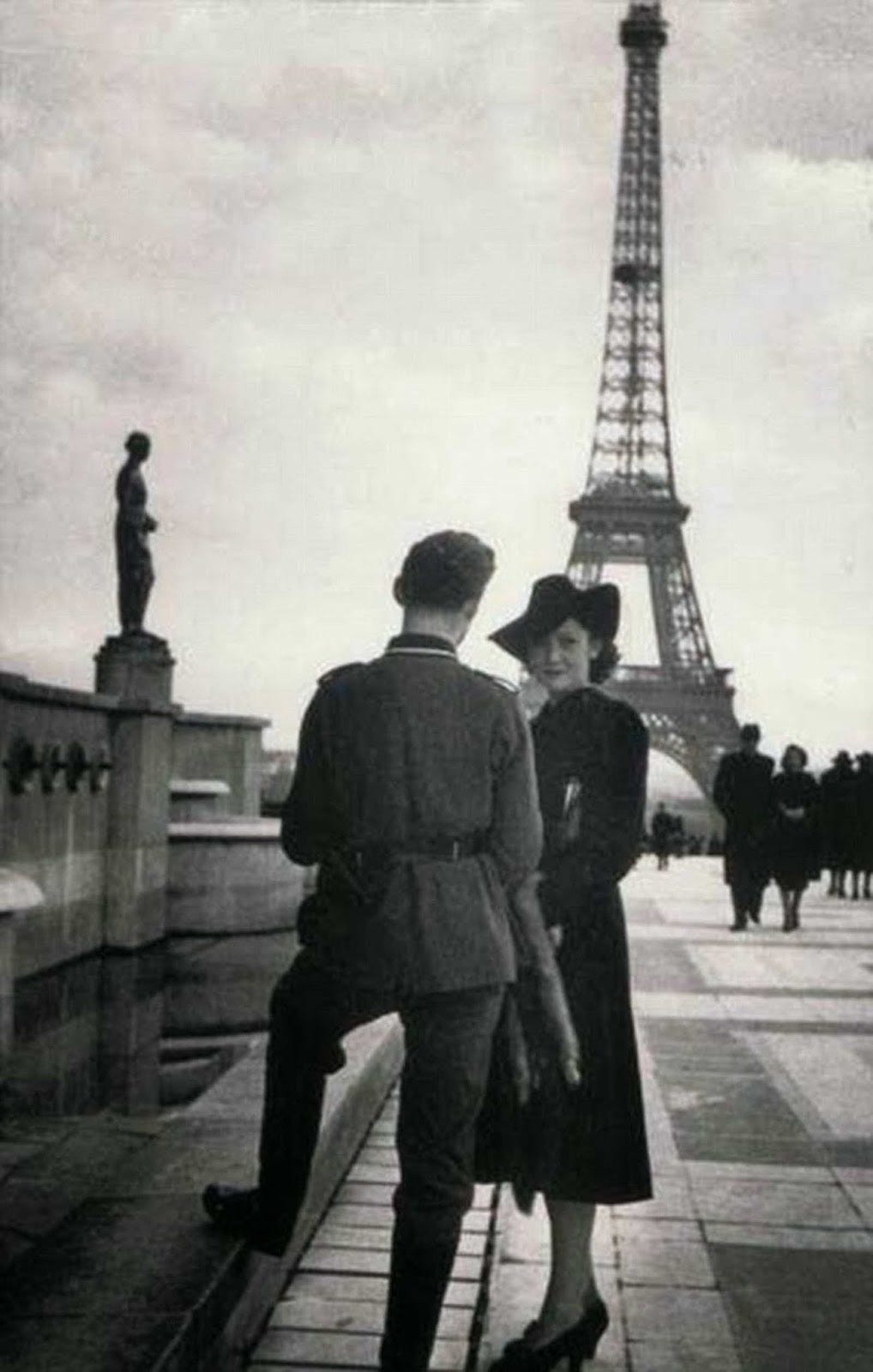 A Frenchwoman, seemingly unperturbed by the occupation, stands with others in the background, challenging assumptions of universal suffering. A Frenchwoman, seemingly unperturbed by the occupation, stands with others in the background, challenging assumptions of universal suffering.
