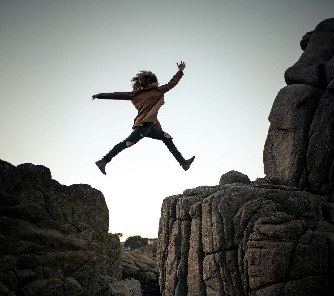 person jumping on big rock under gray and white sky during daytime
