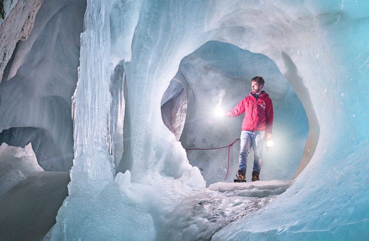 A cave guide in a red jacket stands in front of an impressive ice formation with a carbide lamp and magnesium light. The clear blue ice walls of the cave reveal complex structures, while a taut rope ensures safety.