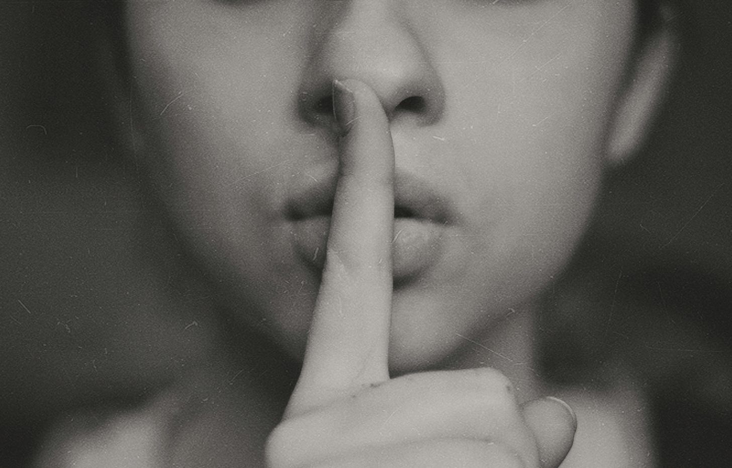 A close-up black-and-white photo of a person with their index finger over their lips, signaling silence. The expression is focused and calm.
