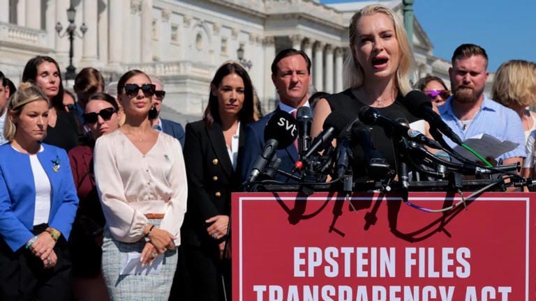 A white woman speaks in front of a podium with a large red sign that says 