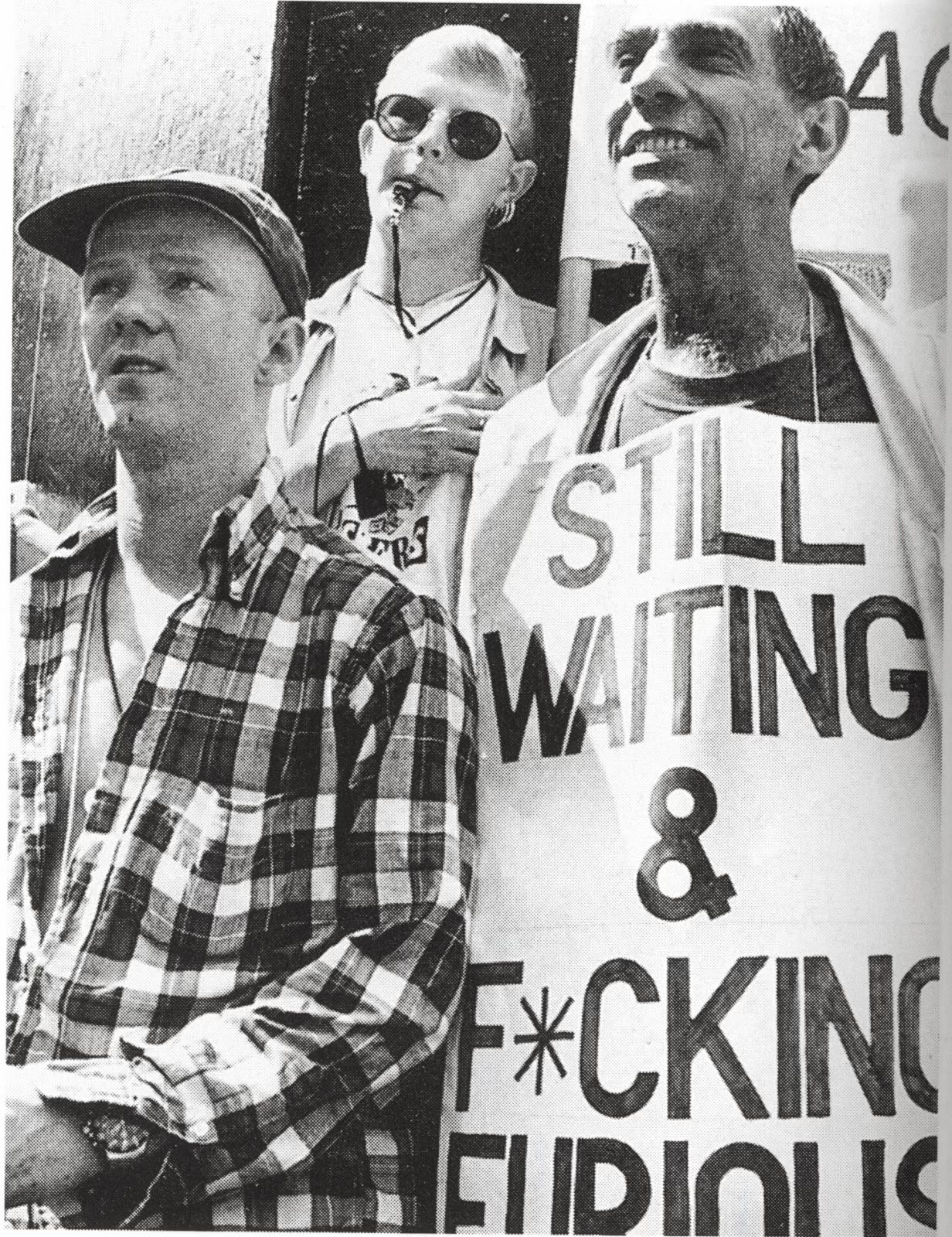 Derek Jarman stands proudly on the right hand side, wearing a banner which reads “STILL WAITING & F*CKING FURIOUS"