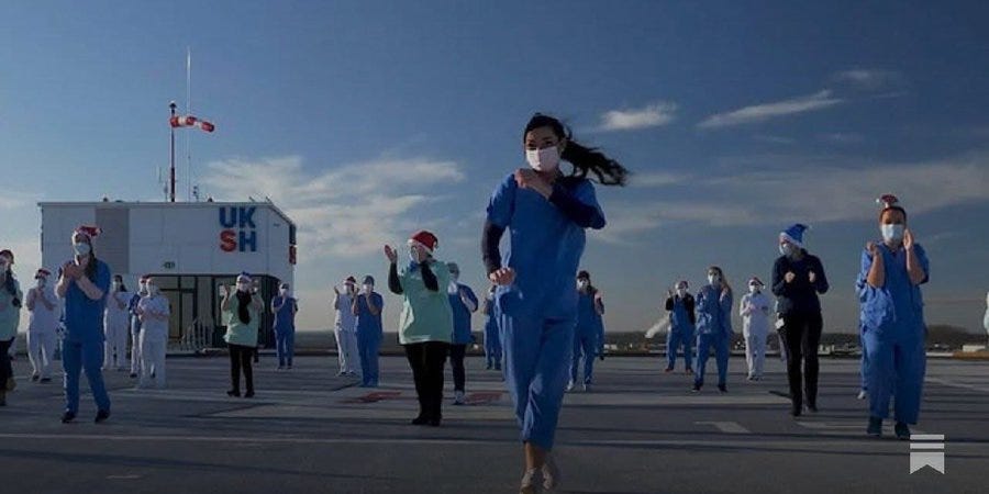 A group of healthcare workers in blue scrubs and white coats, some wearing Santa hats and face masks, stand and run on an outdoor rooftop helipad area next to a building with a Union Jack flag and a sign reading USH. The scene is set under a partly cloudy sky with several individuals clapping and cheering, capturing a celebratory moment among the medical staff. A group of healthcare workers in blue scrubs and white coats, some wearing Santa hats and face masks, stand and run on an outdoor rooftop helipad area next to a building with a Union Jack flag and a sign reading USH. The scene is set under a partly cloudy sky with several individuals clapping and cheering, capturing a celebratory moment among the medical staff.