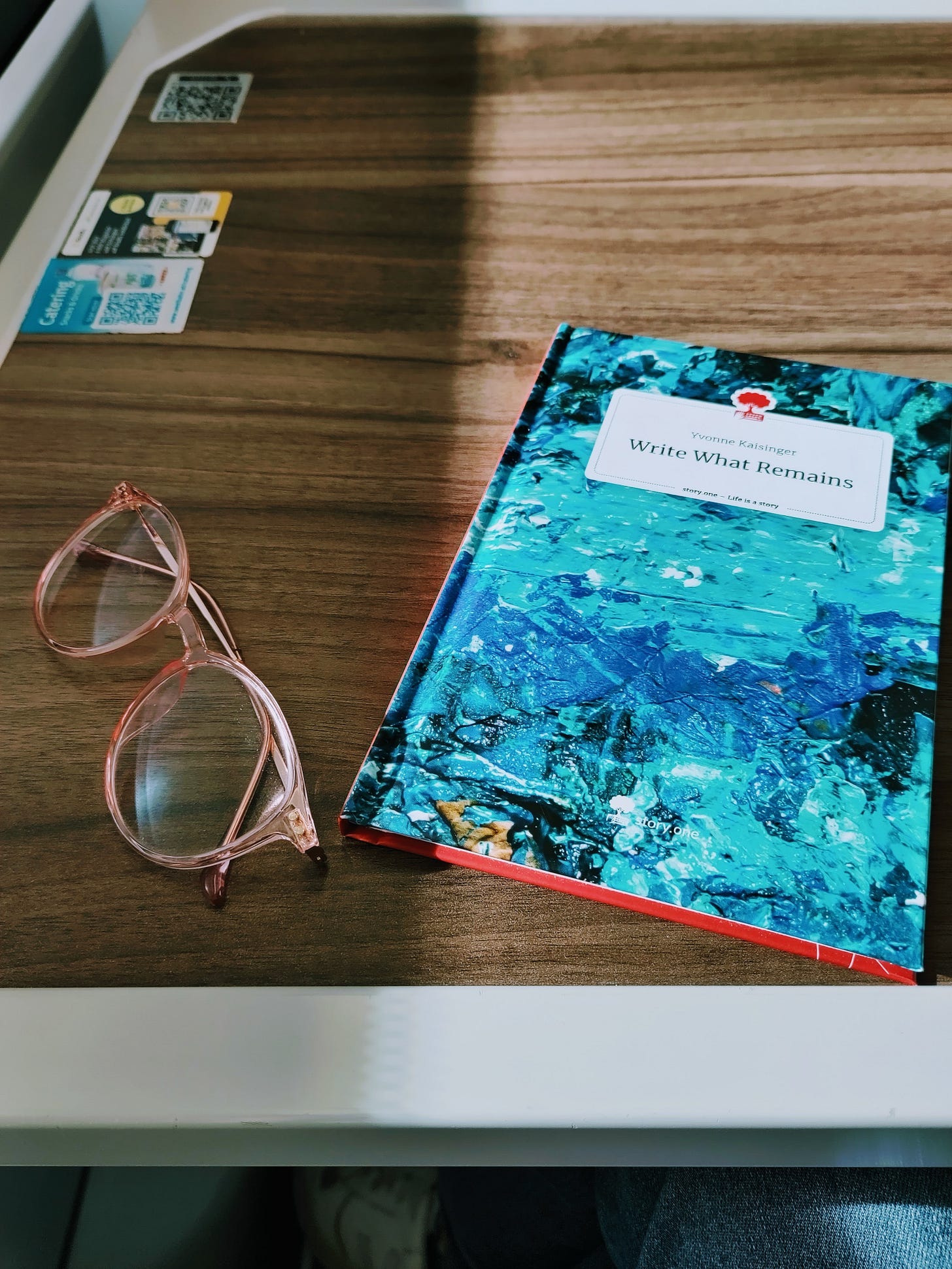 The photo shows the author's published book titled Write What Remains with an artistic, turquoise, blue, and black cover, and her rose glasses on a small table on the train.