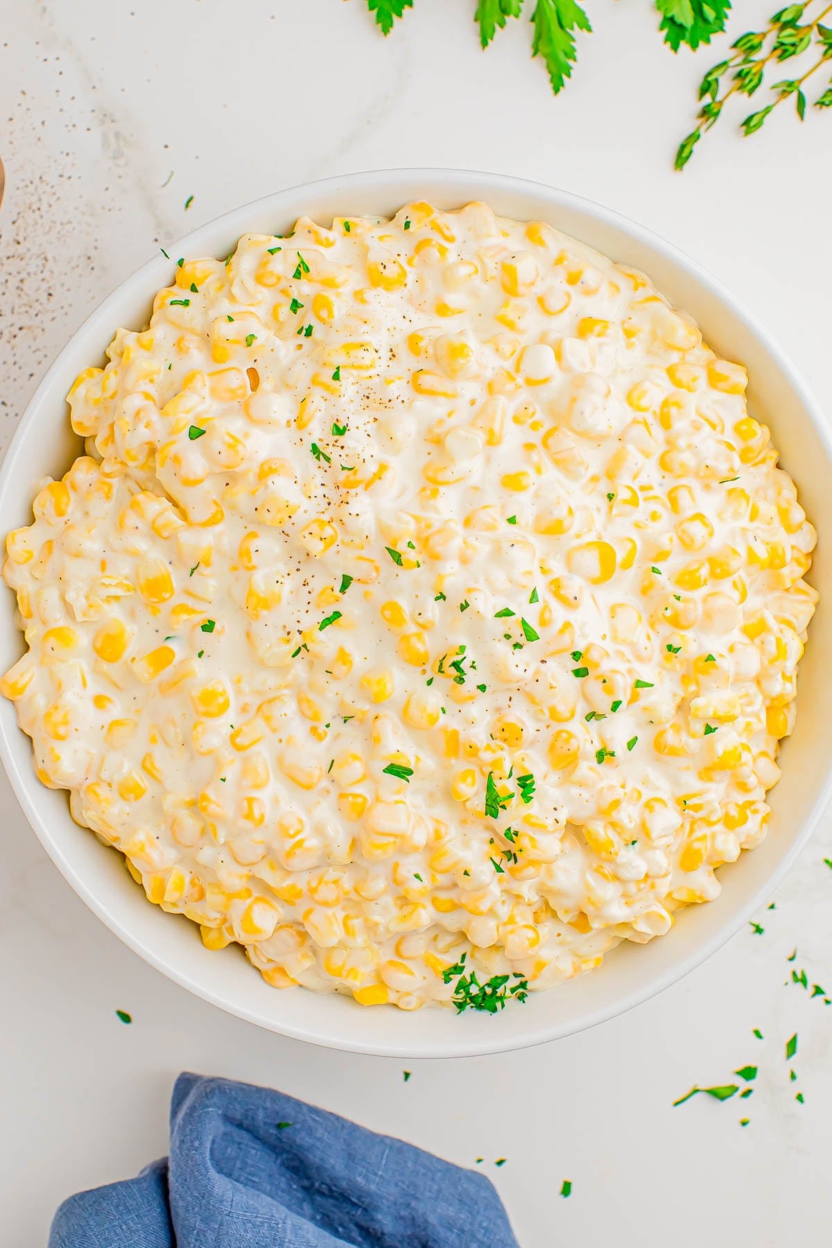 A bowl of creamy corn topped with finely chopped parsley, set on a white surface with a blue napkin and herb sprigs nearby. A bowl of creamy corn topped with finely chopped parsley, set on a white surface with a blue napkin and herb sprigs nearby.