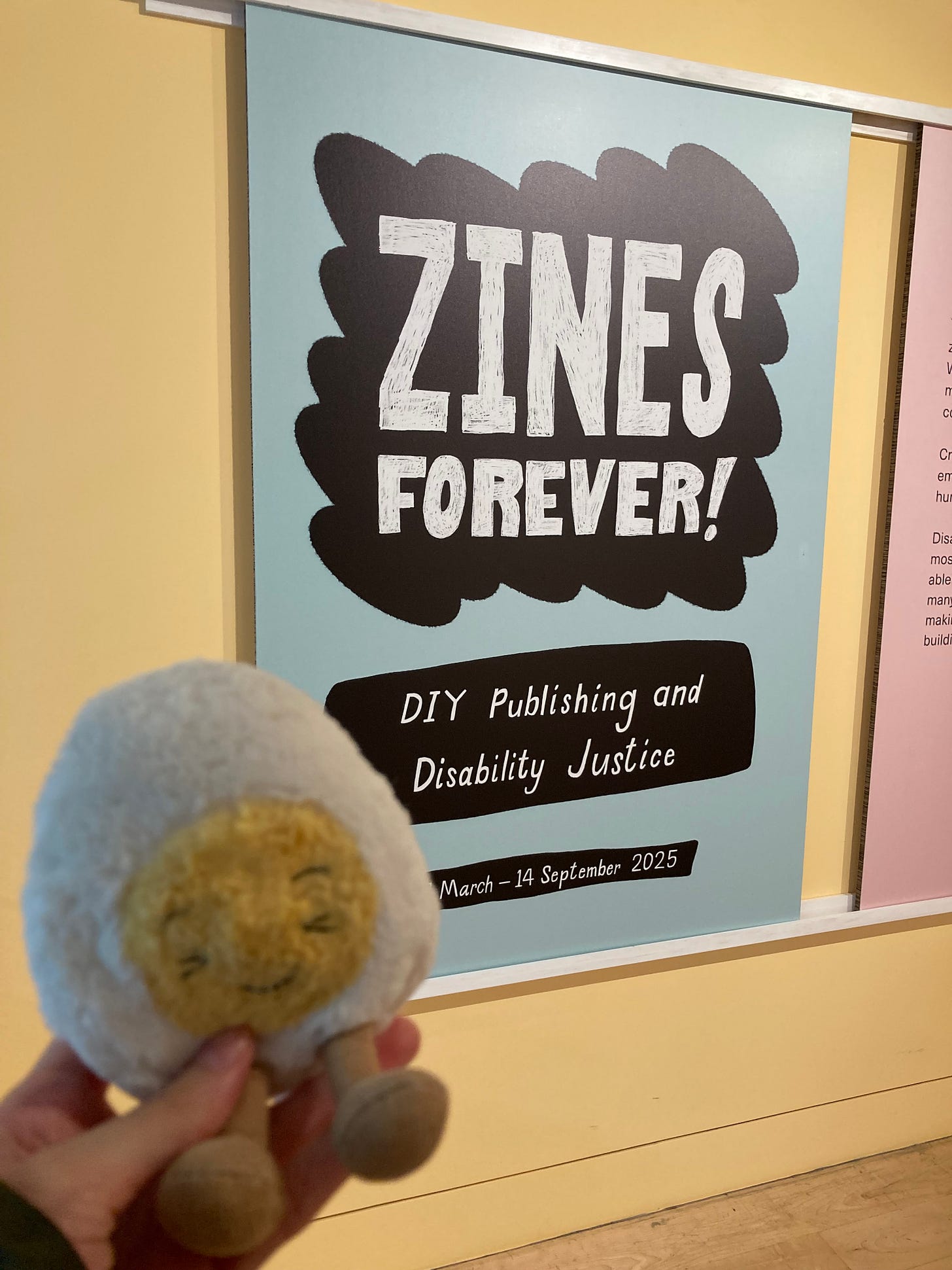 Dippy the soft toy boiled egg in front of a large poster in a museum. The poster reads Zines Forever! DIY publishing and disability justice. 