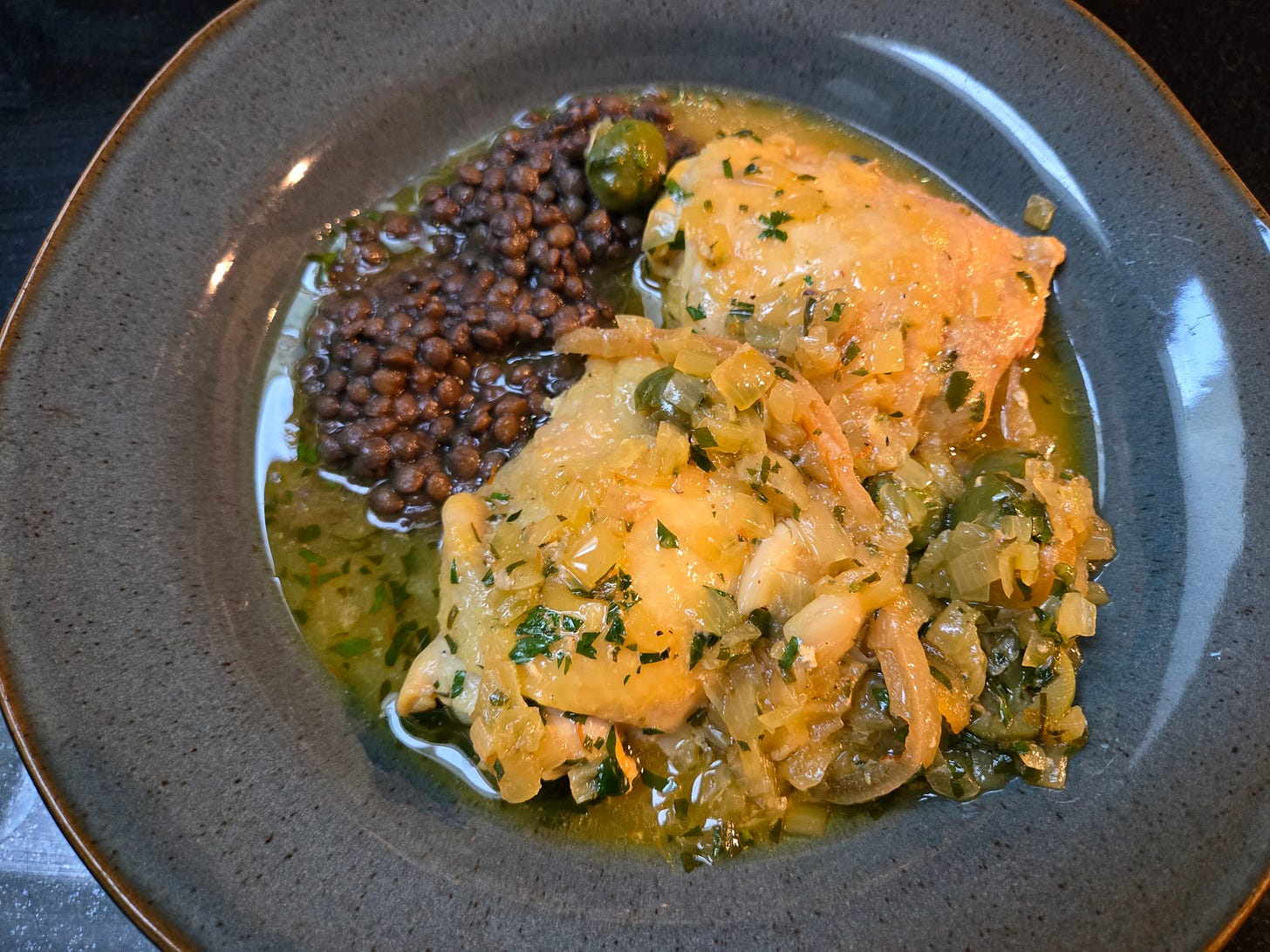 A bowl with two chicken thighs topped with a big pile of preserved lemon strips and olives, a small pile of French lentils, all swimming in tagine liquid. 