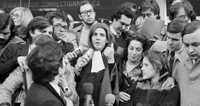 Una foto in bianco e nero: al centro c'è Gisèle Halimi con la toga, è circondata da giornalisti che tendono il microfono verso di lei. Al suo fianco, Marie-Claire Chevalier e un’altra donna che la cinge per le spalle. Una foto in bianco e nero: al centro c'è Gisèle Halimi con la toga, è circondata da giornalisti che tendono il microfono verso di lei. Al suo fianco, Marie-Claire Chevalier e un’altra donna che la cinge per le spalle.
