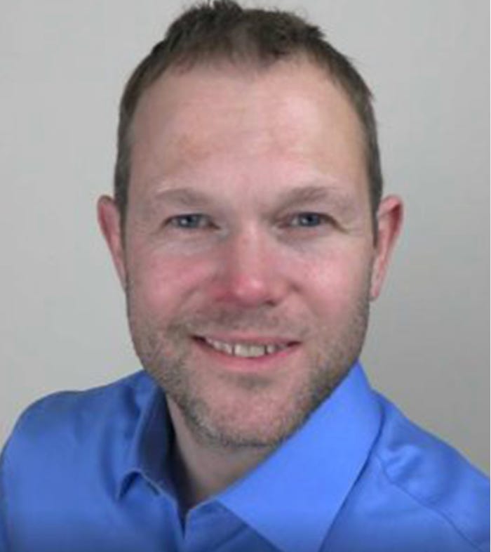 Portrait of Max Kane, a man with short hair and a blue shirt against a plain gray background. Portrait of Max Kane, a man with short hair and a blue shirt against a plain gray background.
