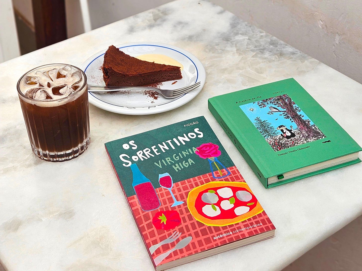 Foto do livro "Os sorrentinos", de Virginia HIga, em uma cafeteria, com um copo de mate gelado e uma torta cheesecake.