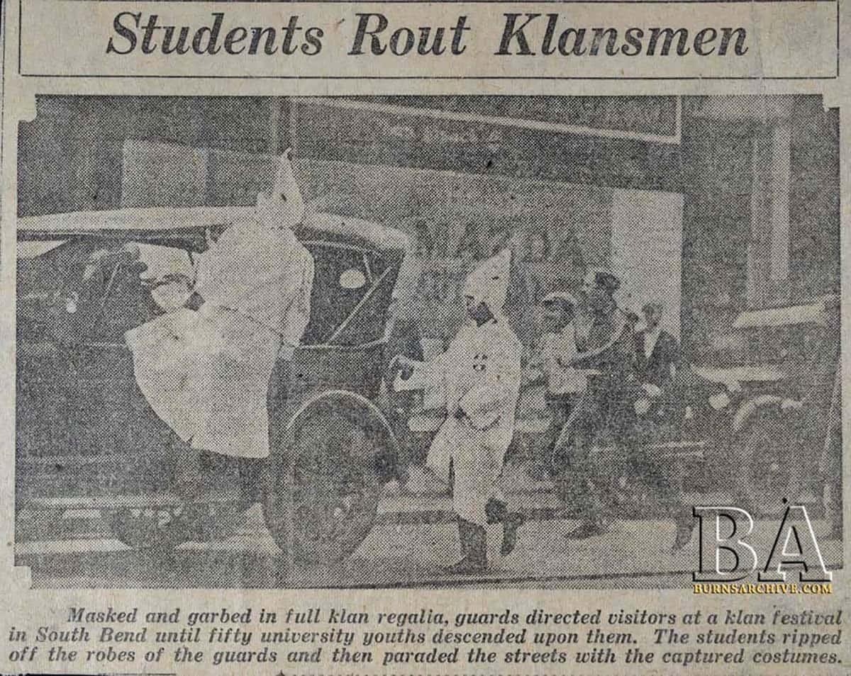 Vintage clipping from a newspaper with the headline 'Students Rout Klansmen', depicting a group of individuals confronting others dressed in Ku Klux Klan regalia next to old cars on a city street. The image captures a moment of confrontation between Notre Dame students and Klan members.