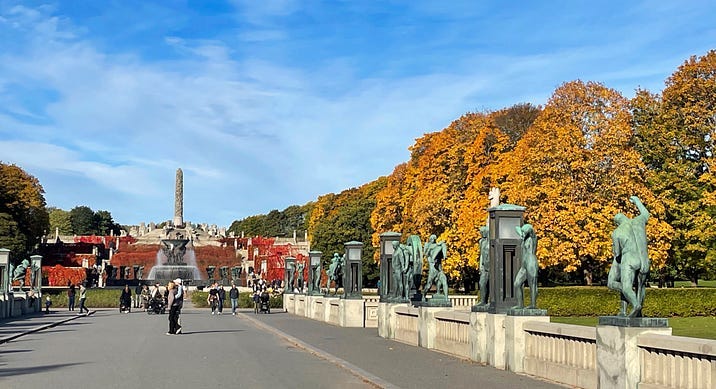 A bridge with statues, a tall monument at the end, colorful autumn trees on the background.