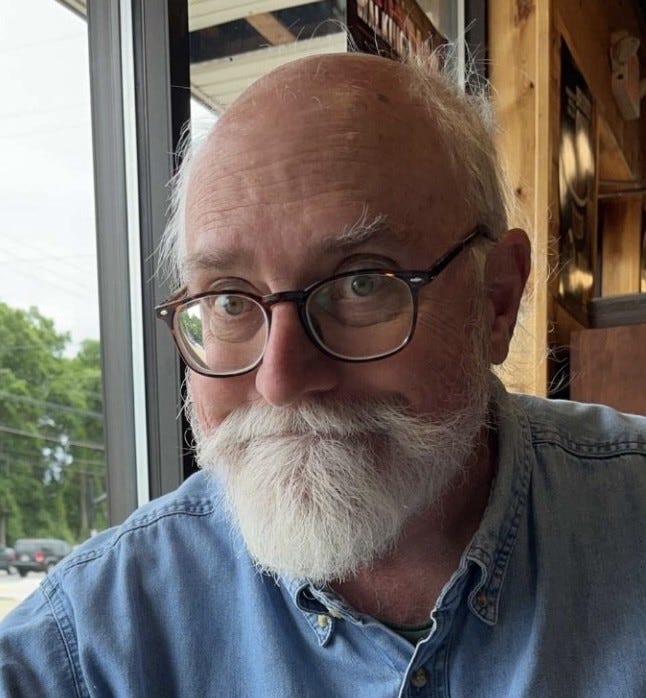 A close-up portrait shows an older man with a full white beard and round glasses seated indoors near a large window, natural light illuminating his face. He wears a denim button-down shirt and looks off-camera with a calm, reflective expression. The warm wood interior and soft daylight create a quiet, contemplative mood suggestive of conversation or reflection. A close-up portrait shows an older man with a full white beard and round glasses seated indoors near a large window, natural light illuminating his face. He wears a denim button-down shirt and looks off-camera with a calm, reflective expression. The warm wood interior and soft daylight create a quiet, contemplative mood suggestive of conversation or reflection.