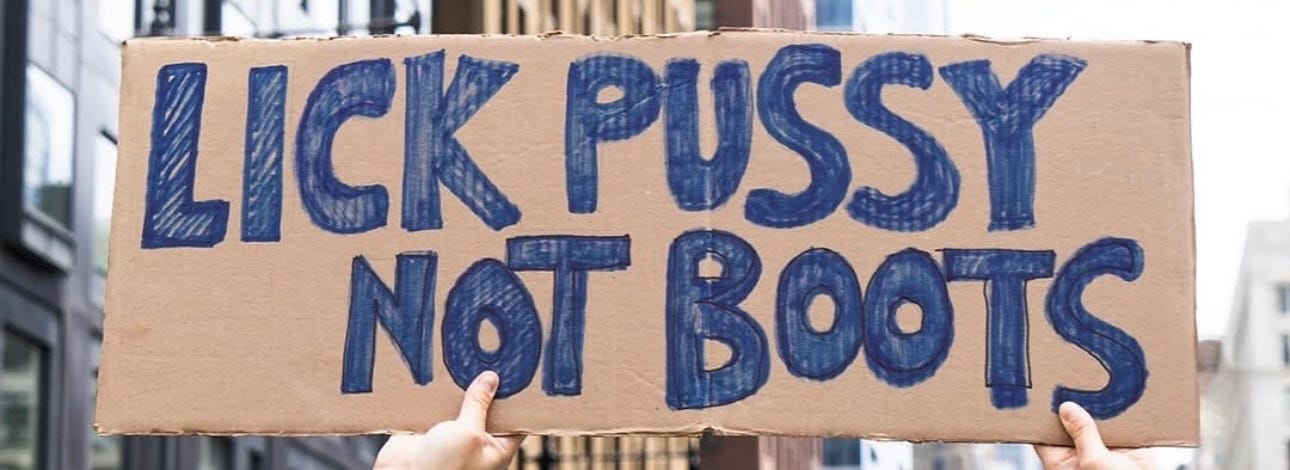 A cardboard protest sign that reads LICK PUSSY NOT BOOTS