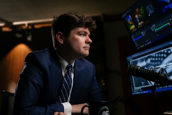 A young white man in a blue suit in front of a microphone and computer screen. A young white man in a blue suit in front of a microphone and computer screen.