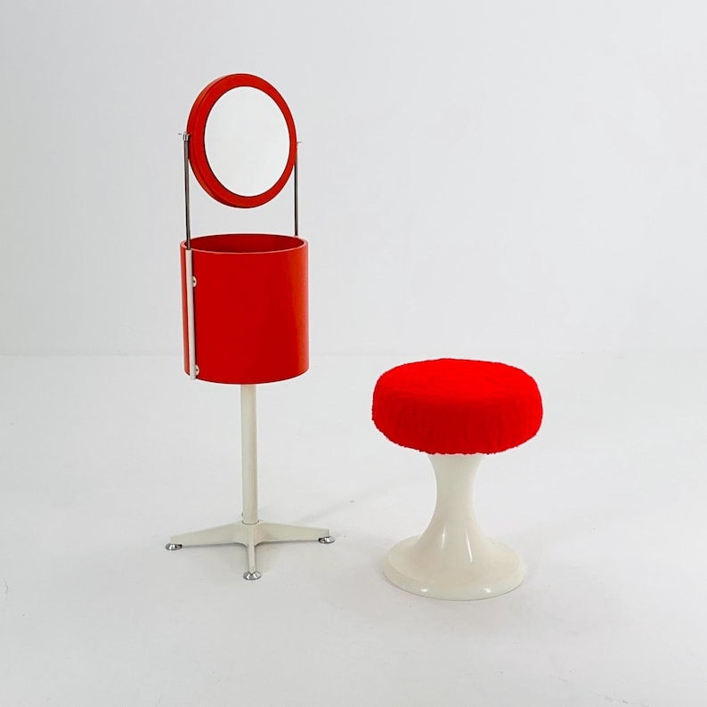 May include: A red vanity set with a round mirror and a white base. The vanity has a red cylindrical storage compartment. A red and white stool with a white base is next to the vanity.