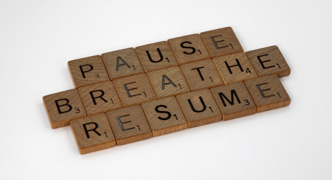On the white background, three lines stacked of wooden Scrabble blocks that spell PAUSE, BREATHE, RESUME. 