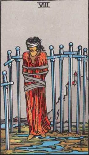 imagem da carta de tarot 8 de espadas onde se vê uma mulher amarrada, vendada, cercada por trás por espadas enfiadas no chão de terra, ao fundo se vê uma montanha com um castelo no topo