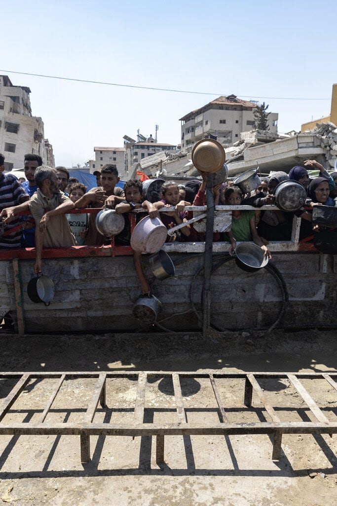 Palestinians holding metal and plastic bowls crowd against a wall, awaiting food distribution.