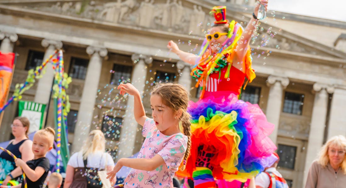 A girl wearing a pink t-shirt dancing, with a crowd of people and a stilt walker behind her A girl wearing a pink t-shirt dancing, with a crowd of people and a stilt walker behind her
